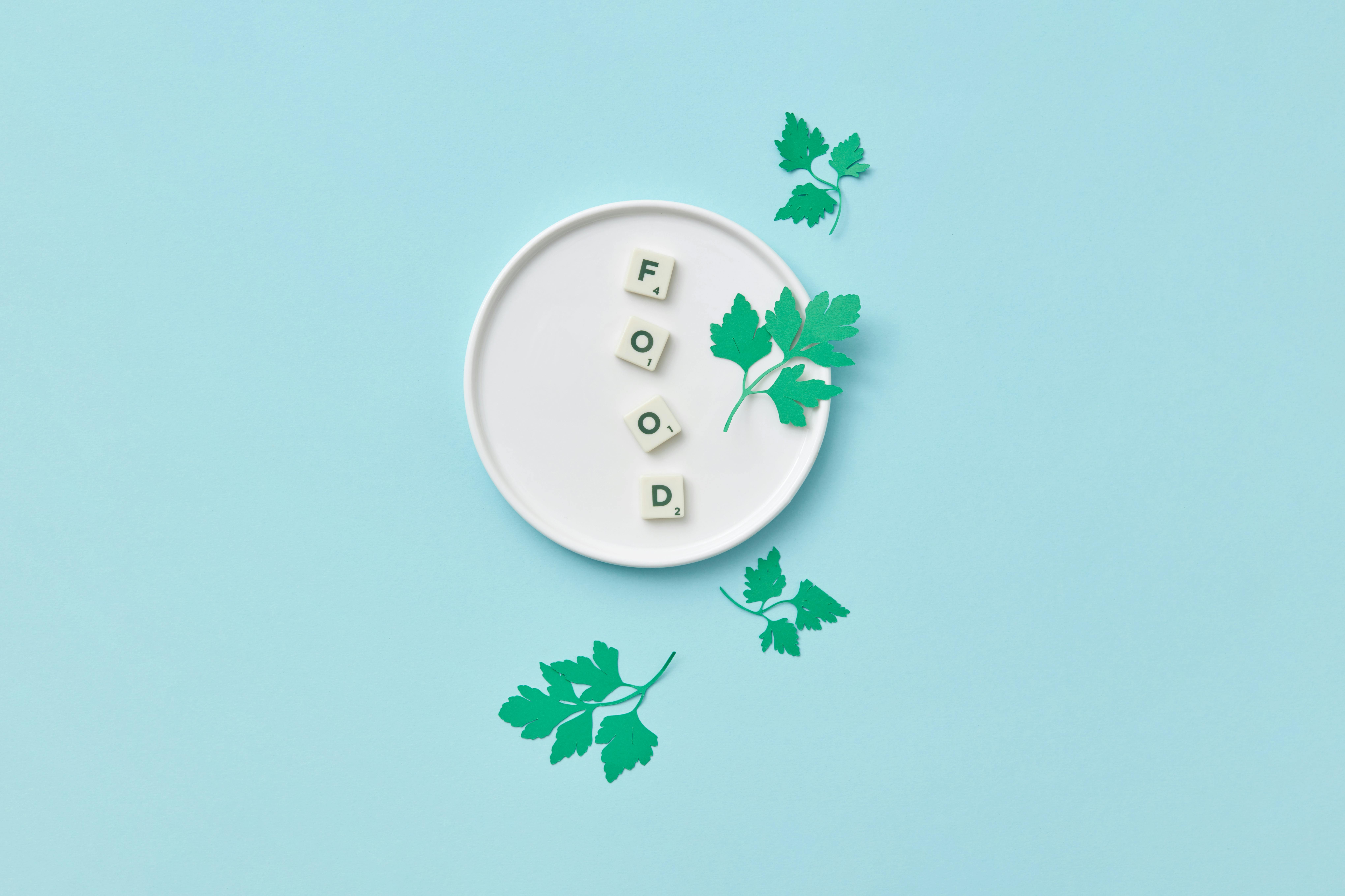 A minimalist composition featuring Scrabble tiles spelling 'food' with parsley on a white plate against a blue background.